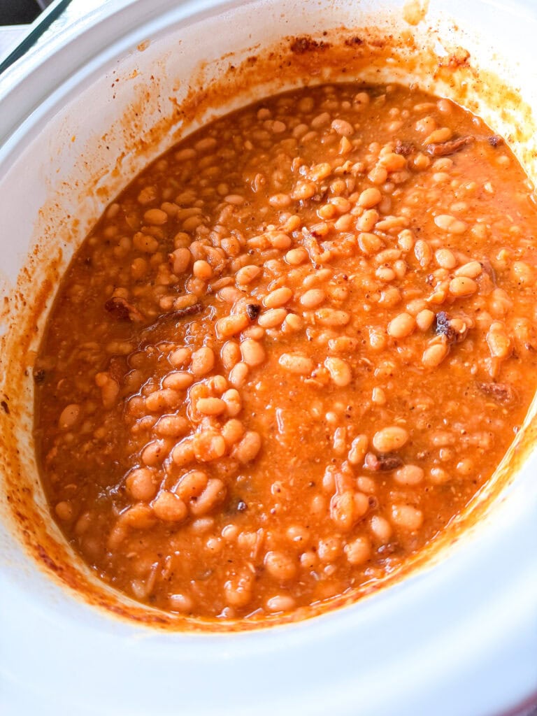 A close-up of baked beans in a white slow cooker, showing a thick, orange-brown sauce with visible beans and bits of meat.