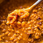 A close-up of a wooden spoon holding baked beans in a thick, rich orange sauce, with more beans visible in the pot below.