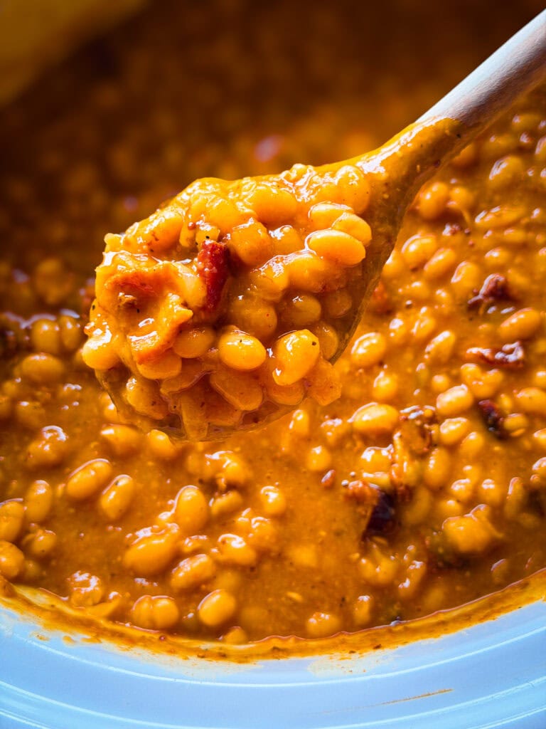 A close-up of a wooden spoon holding baked beans in a thick, rich orange sauce, with more beans visible in the pot below.