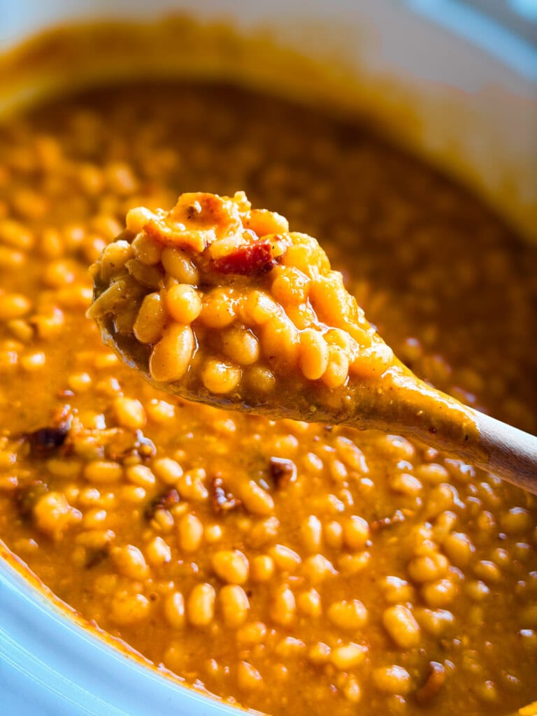 A close-up of a wooden spoon lifting baked beans in a rich, thick sauce from a pot. The beans appear tender and saucy, with visible bits of bacon or seasoning mixed in.