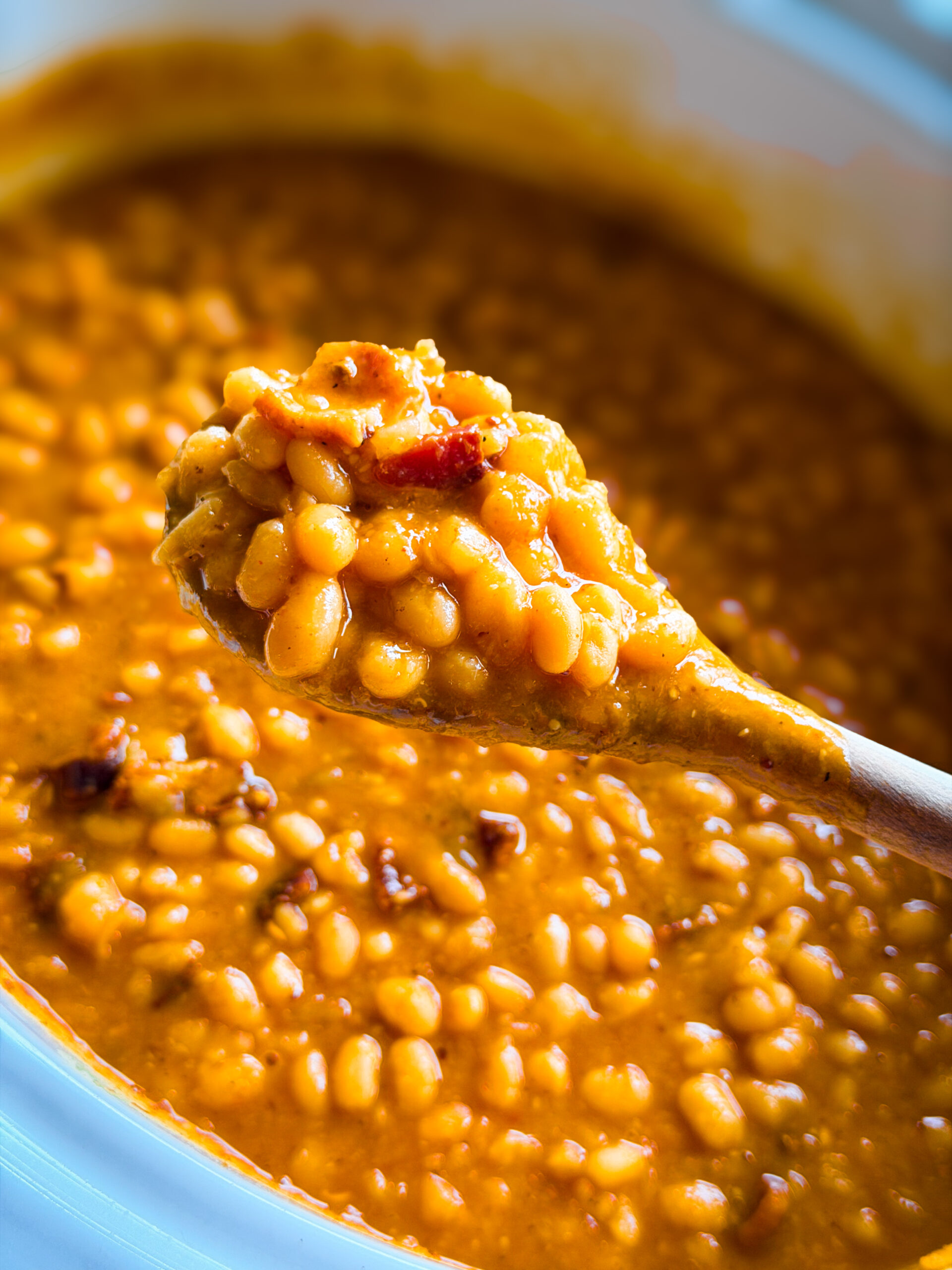 A close-up of a wooden spoon lifting baked beans in a rich, thick sauce from a pot. The beans appear tender and saucy, with visible bits of bacon or seasoning mixed in.