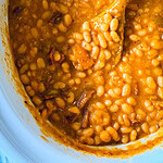 A close-up of baked beans in a thick, orange-brown sauce being stirred with a wooden spoon inside a white pot.