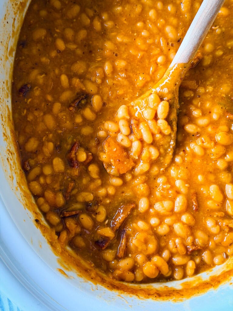 A close-up of baked beans in a thick, orange-brown sauce being stirred with a wooden spoon inside a white pot.