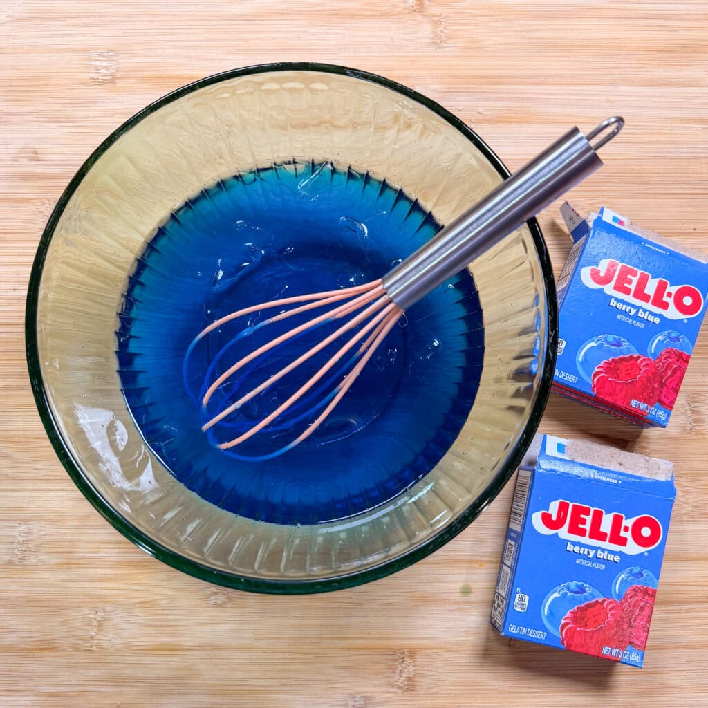 A glass bowl with blue Jello being whisked sits on a wooden surface. Next to the bowl are two boxes of berry blue Jello mix.