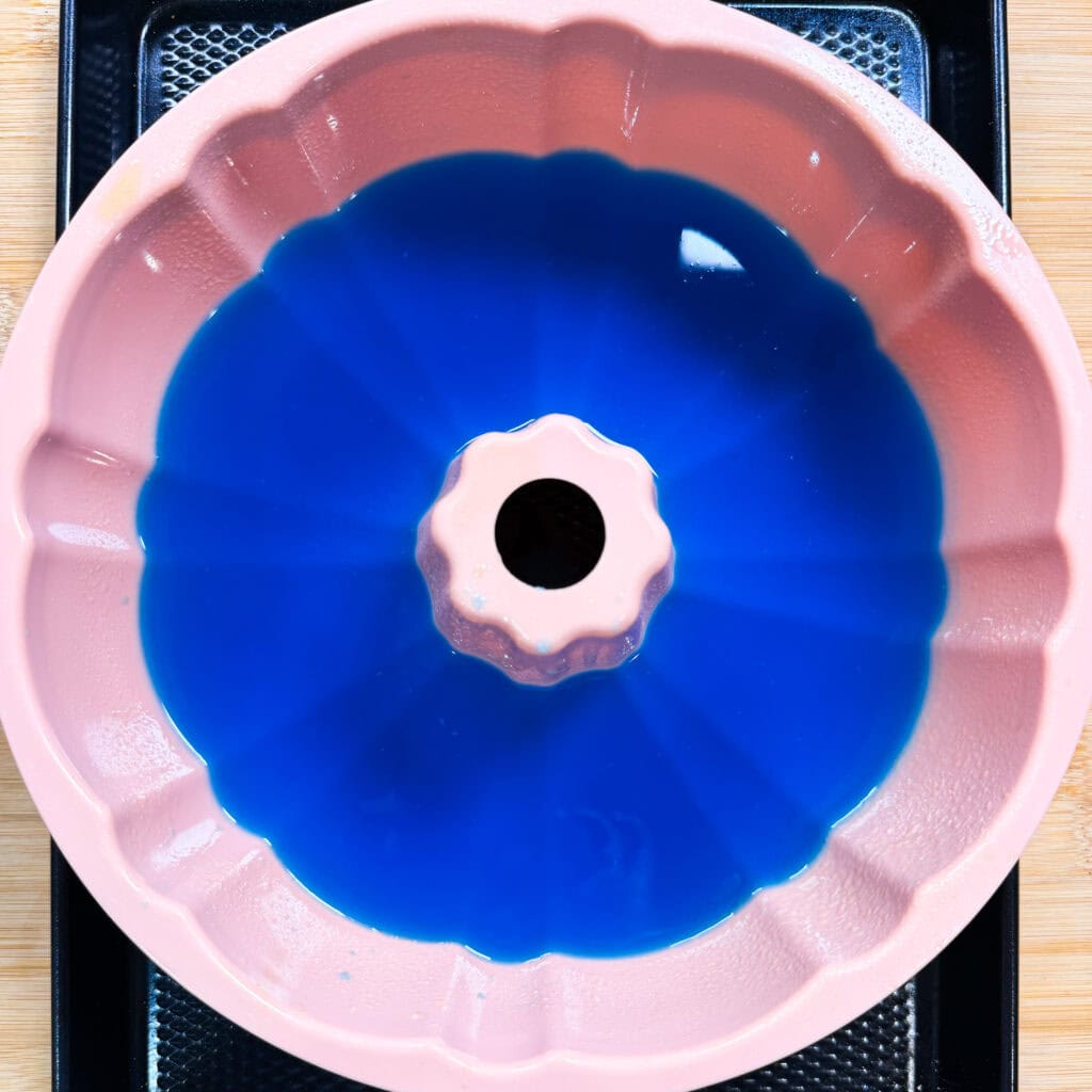 A pink bundt cake pan filled with blue liquid, placed on a black tray over a wooden surface, viewed from above.