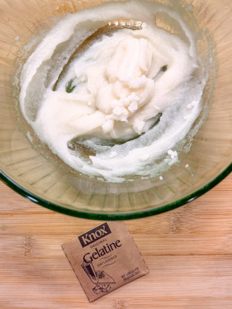 A glass bowl contains a white, grainy mixture, likely sugar and gelatin, on a wooden surface. In front of the bowl is a packet labeled Knox Original Gelatine Unflavored.