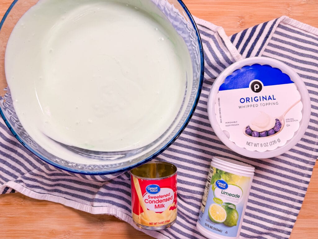 A bowl of light green creamy mixture sits on a striped cloth next to containers of whipped topping, frozen limeade, and a can of sweetened condensed milk on a wooden surface.