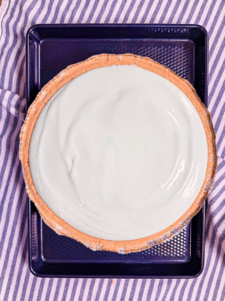 A graham cracker pie crust filled with smooth, pale green mint pie filling sits on a dark baking tray atop a striped cloth.