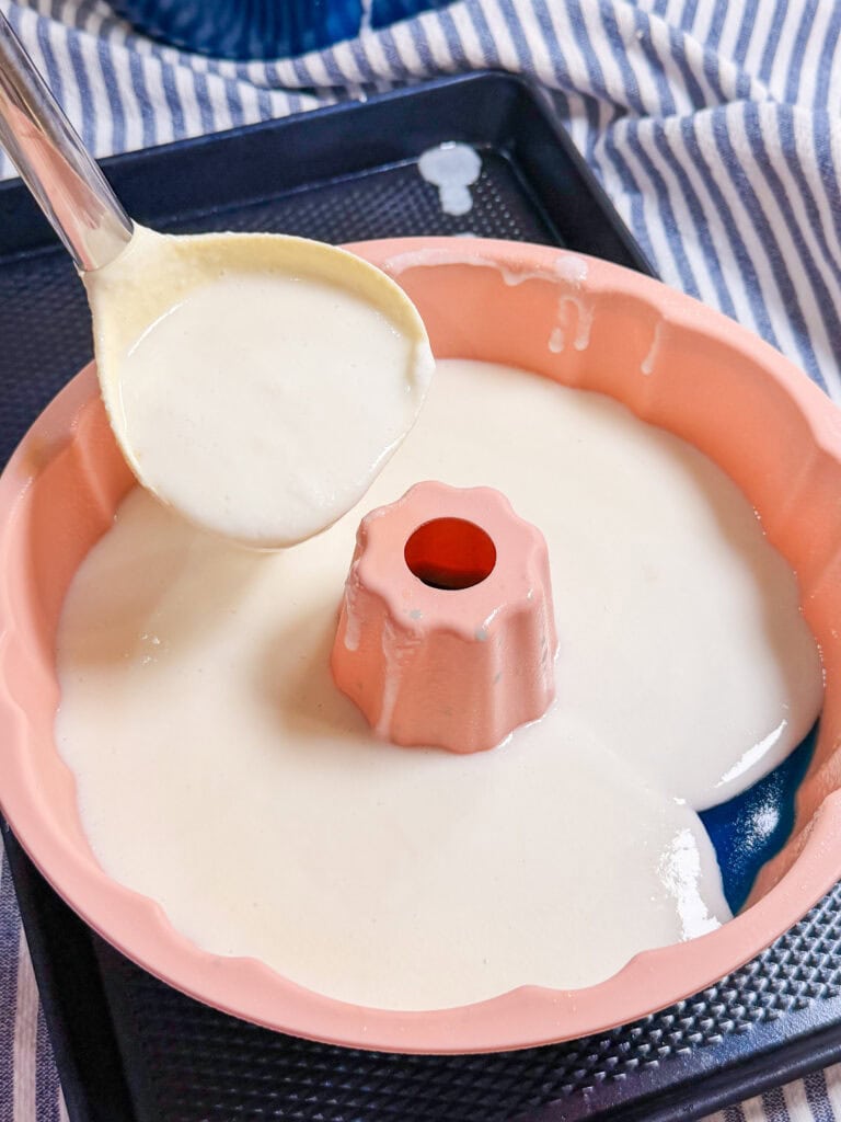 A close-up of cake batter being poured from a ladle into a pink Bundt cake pan, which sits on a black tray with a striped cloth in the background.