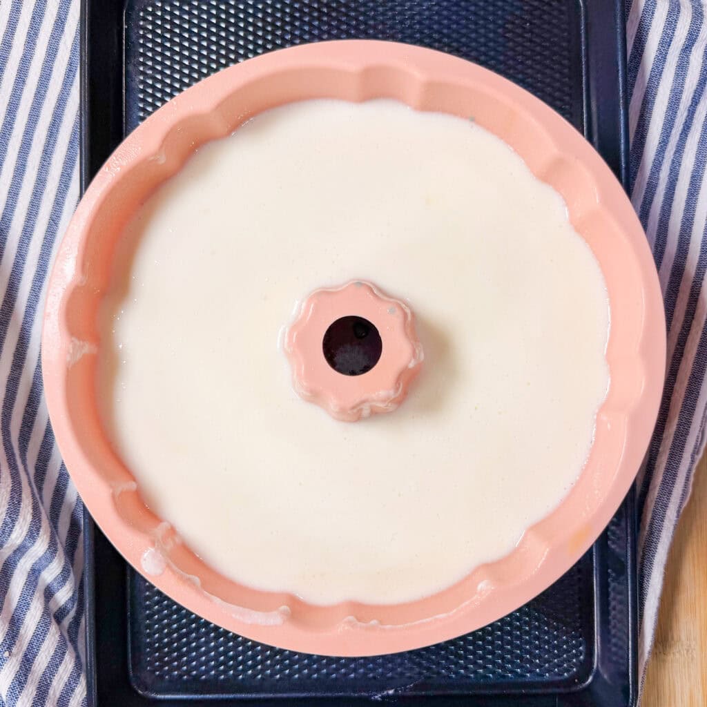 A pink bundt cake pan filled with white batter sits on a black baking tray, with a blue and white striped cloth partially visible beside it.