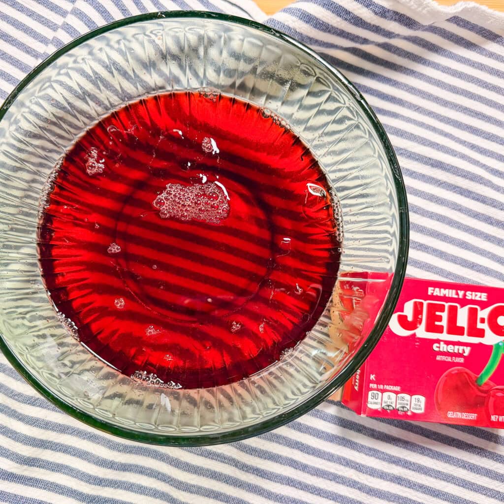 A glass bowl filled with red liquid, likely cherry Jell-O, sits on a blue and white striped cloth next to a family-size box of cherry Jell-O.