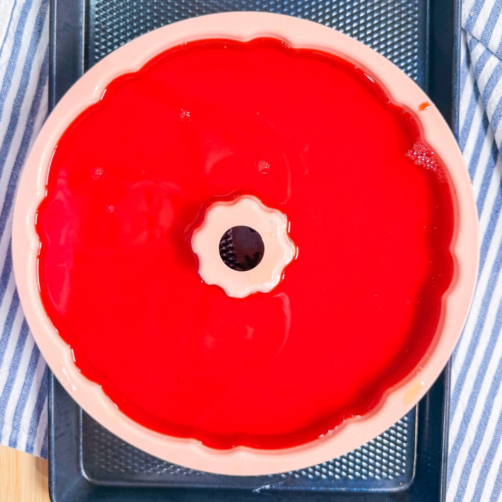 A top-down view of a bundt pan filled with bright red gelatin, sitting on a black tray with a blue and white striped cloth on the side.