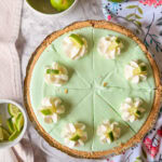 A whole key lime pie with a graham cracker crust, topped with whipped cream and lime wedges, sits on a marble surface next to lime slices in bowls and a colorful floral cloth.