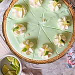 A key lime pie topped with eight dollops of whipped cream, each garnished with a small wedge of lime, is sliced and ready to serve. Sliced limes and a floral-patterned cloth are nearby.