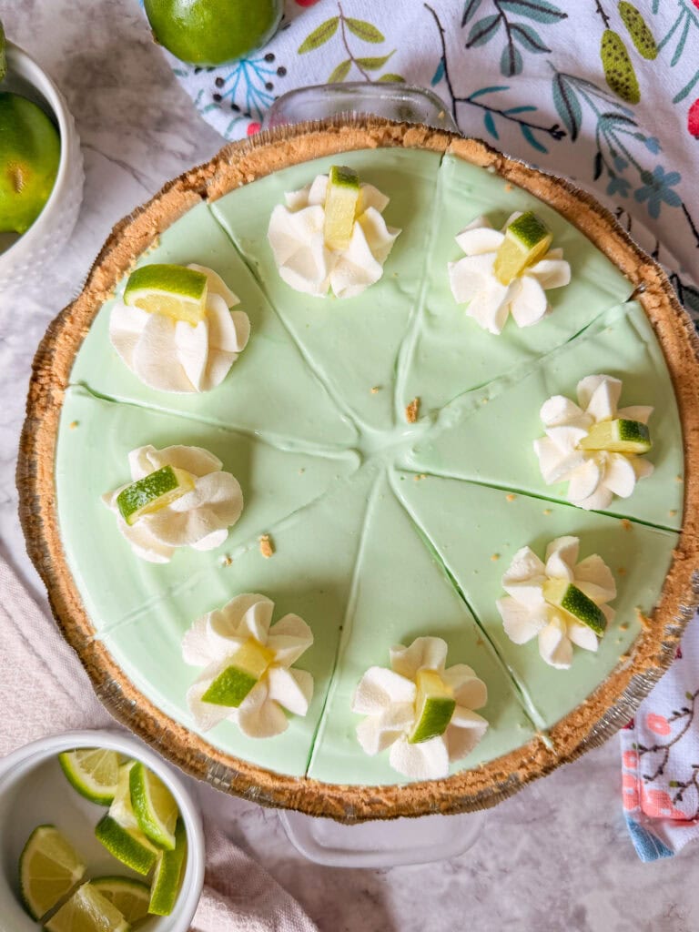 A key lime pie topped with eight dollops of whipped cream, each garnished with a small wedge of lime, is sliced and ready to serve. Sliced limes and a floral-patterned cloth are nearby.