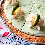 A creamy light green pie with a crumbly crust, topped with whipped cream, lime wedges, and slices, cut into pieces. A cup with lime wedges is in the background, and a colorful towel is partly visible.