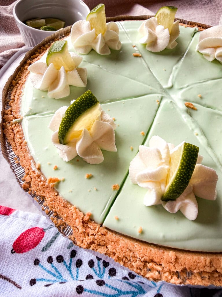 A creamy light green pie with a crumbly crust, topped with whipped cream, lime wedges, and slices, cut into pieces. A cup with lime wedges is in the background, and a colorful towel is partly visible.