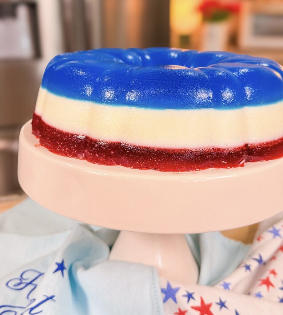 A layered gelatin dessert in blue, white, and red colors sits on a white cake stand, with a patriotic-themed cloth underneath. The background is a kitchen setting.