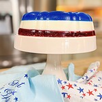 A layered gelatin dessert in red, white, and blue sits on a white cake stand. A star-patterned cloth is draped nearby, evoking a patriotic or festive theme. The background is softly blurred.