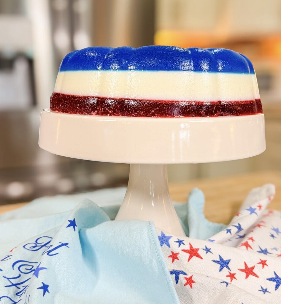 A layered gelatin dessert in red, white, and blue sits on a white cake stand. A star-patterned cloth is draped nearby, evoking a patriotic or festive theme. The background is softly blurred.
