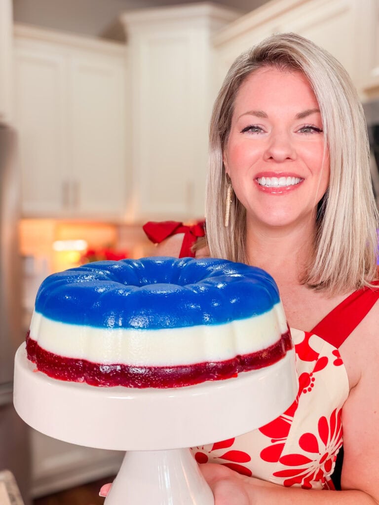 A smiling woman in a red and white floral dress holds a layered gelatin dessert with blue, white, and red sections on a cake stand in a bright kitchen.