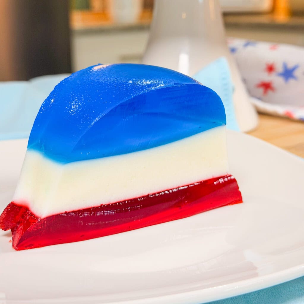 A slice of layered gelatin dessert with red, white, and blue layers sits on a white plate, suggesting a patriotic or festive theme.