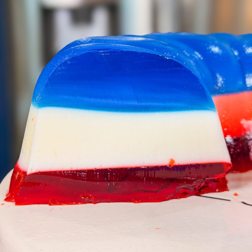 A close-up of a slice of layered gelatin dessert with three distinct colors: red at the bottom, white in the middle, and blue on top, placed on a white surface.