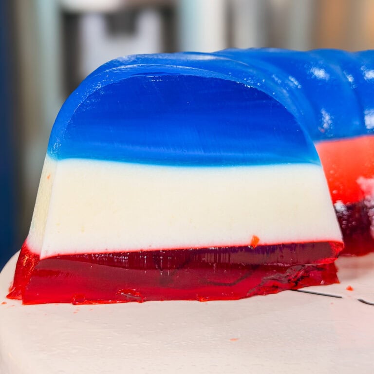 A close-up of a slice of layered gelatin dessert with three distinct colors: red at the bottom, white in the middle, and blue on top, placed on a white surface.