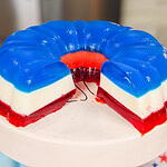 A layered gelatin dessert in red, white, and blue sits on a white cake stand. A large wedge has been cut out, revealing the colorful layers. A star-patterned cloth is nearby.
