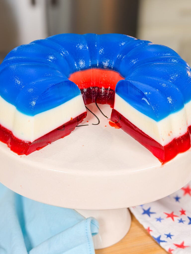 A layered gelatin dessert in red, white, and blue sits on a white cake stand. A large wedge has been cut out, revealing the colorful layers. A star-patterned cloth is nearby.