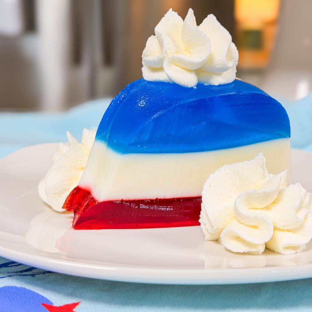 A slice of layered gelatin dessert with red, white, and blue layers, topped and garnished with swirls of whipped cream, on a white plate.
