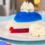 A slice of layered gelatin dessert with red, white, and blue layers, topped and garnished with swirls of whipped cream, on a white plate.