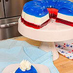 A slice of layered red, white, and blue gelatin dessert topped with whipped cream sits on a plate, with the rest of the gelatin mold on a cake stand in the background. Patriotic-themed décor is visible.