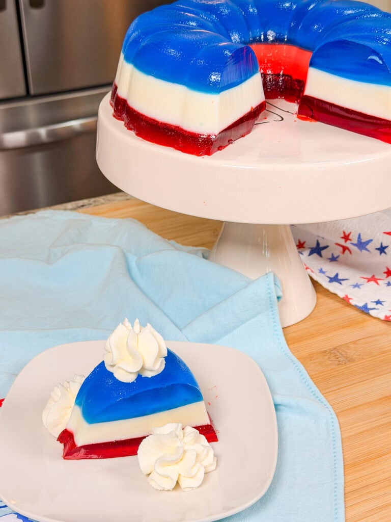 A slice of layered red, white, and blue gelatin dessert topped with whipped cream sits on a plate, with the rest of the gelatin mold on a cake stand in the background. Patriotic-themed décor is visible.