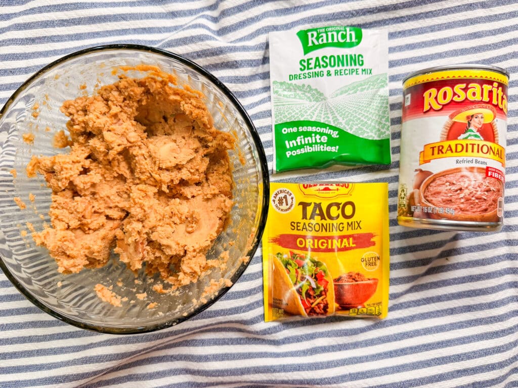 A glass bowl with refried beans sits on a striped cloth next to a can of Rosarita refried beans, a packet of ranch seasoning, and a packet of taco seasoning mix.