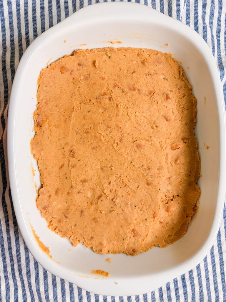 A white baking dish filled with an even layer of orange bean mixture, set on a blue and white striped cloth.