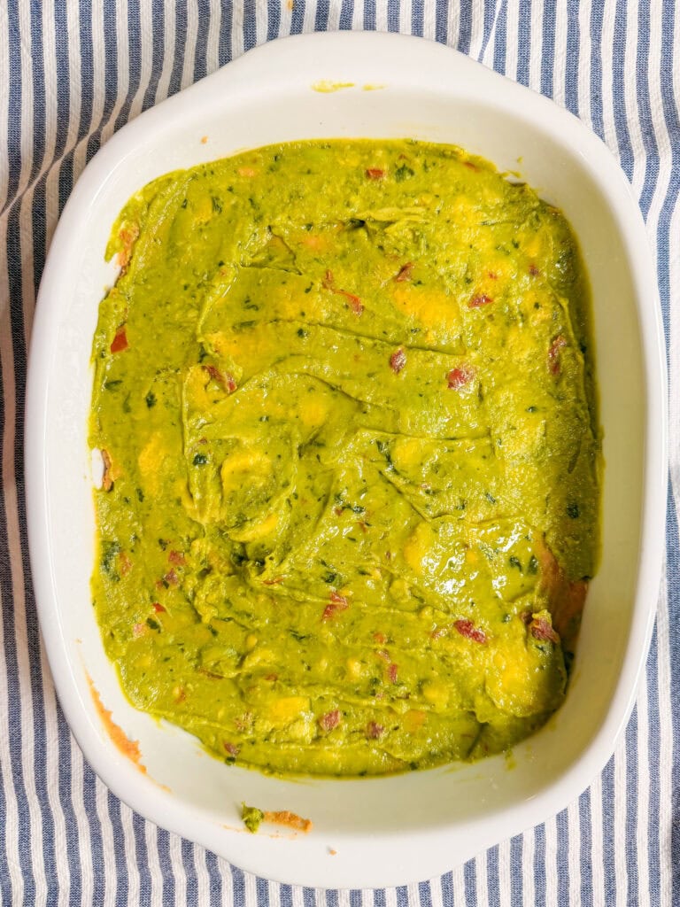 A white baking dish filled with creamy, chunky guacamole, featuring visible pieces of avocado, tomato, and herbs, sits on a blue and white striped cloth.