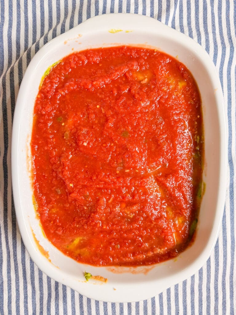 A white oval dish filled with a layer of red tomato sauce, placed on a blue and white striped fabric background.