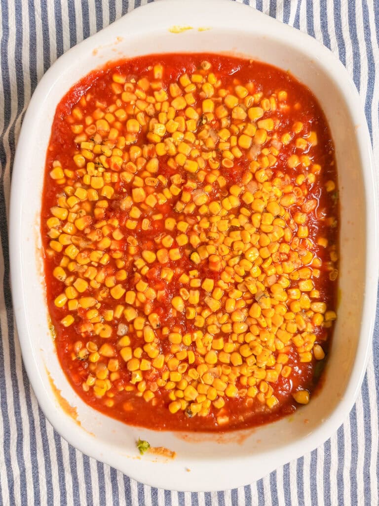 A white baking dish filled with tomato sauce and topped with a layer of yellow corn kernels, set on a blue-and-white striped cloth.