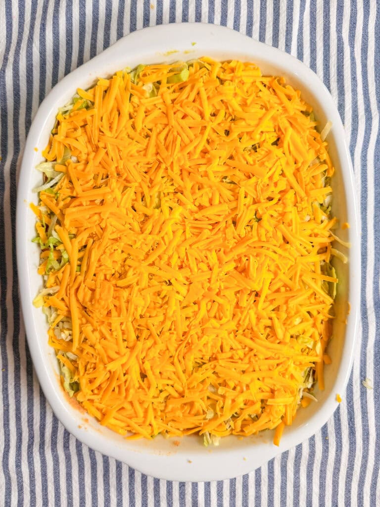 A white baking dish filled with a casserole topped with a generous layer of shredded cheddar cheese, placed on a blue and white striped cloth.