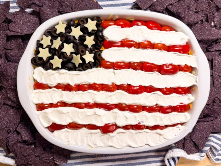 A dip tray decorated like the American flag, with black olives and star-shaped cheese for the stars, cherry tomatoes for the red stripes, and a creamy white layer for the white stripes. Blue corn chips surround the tray.