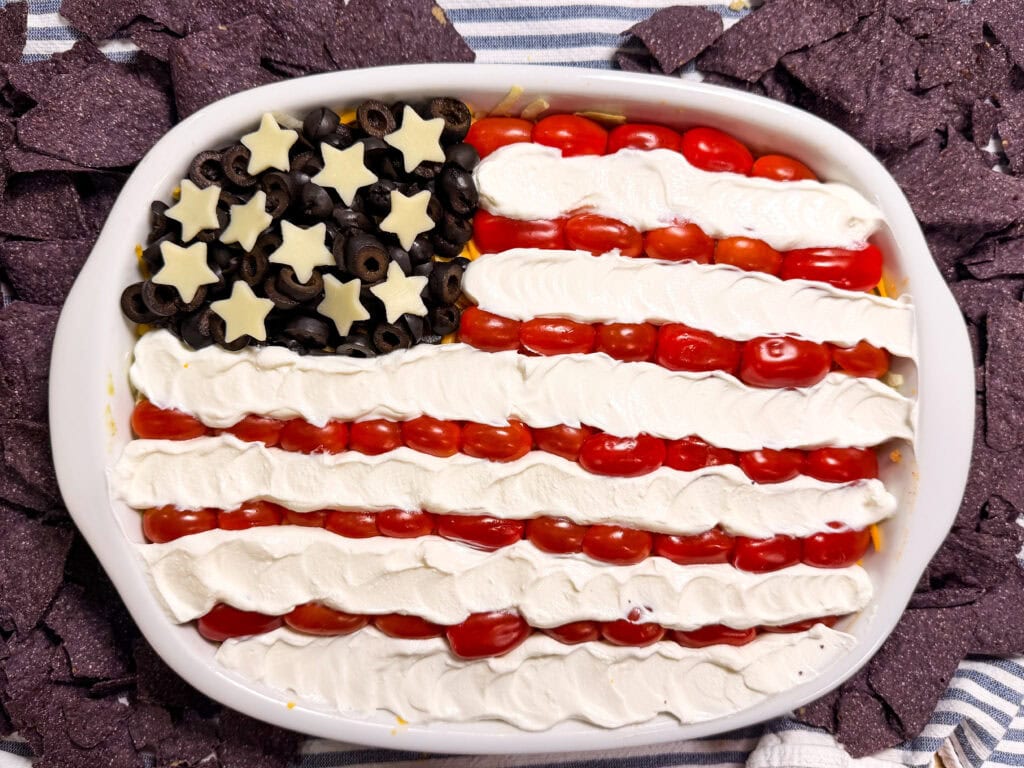 A dip arranged to look like the American flag, with black olives and cheese stars for the blue field, rows of cherry tomatoes for red stripes, and sour cream for white stripes, surrounded by blue corn tortilla chips.
