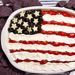 A dip arranged to look like the American flag, with black olives and cheese stars for the blue field, rows of cherry tomatoes for red stripes, and sour cream for white stripes, surrounded by blue corn tortilla chips.
