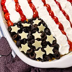 A layered dip arranged to look like the American flag, with rows of cherry tomatoes and sour cream for stripes, and chopped black olives with star-shaped cheese pieces for the stars on a white oval dish.