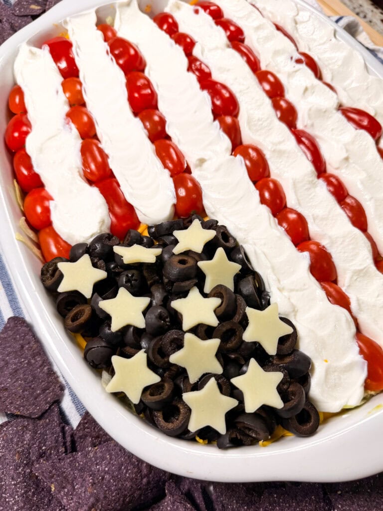 A layered dip arranged to look like the American flag, with rows of cherry tomatoes and sour cream for stripes, and chopped black olives with star-shaped cheese pieces for the stars on a white oval dish.