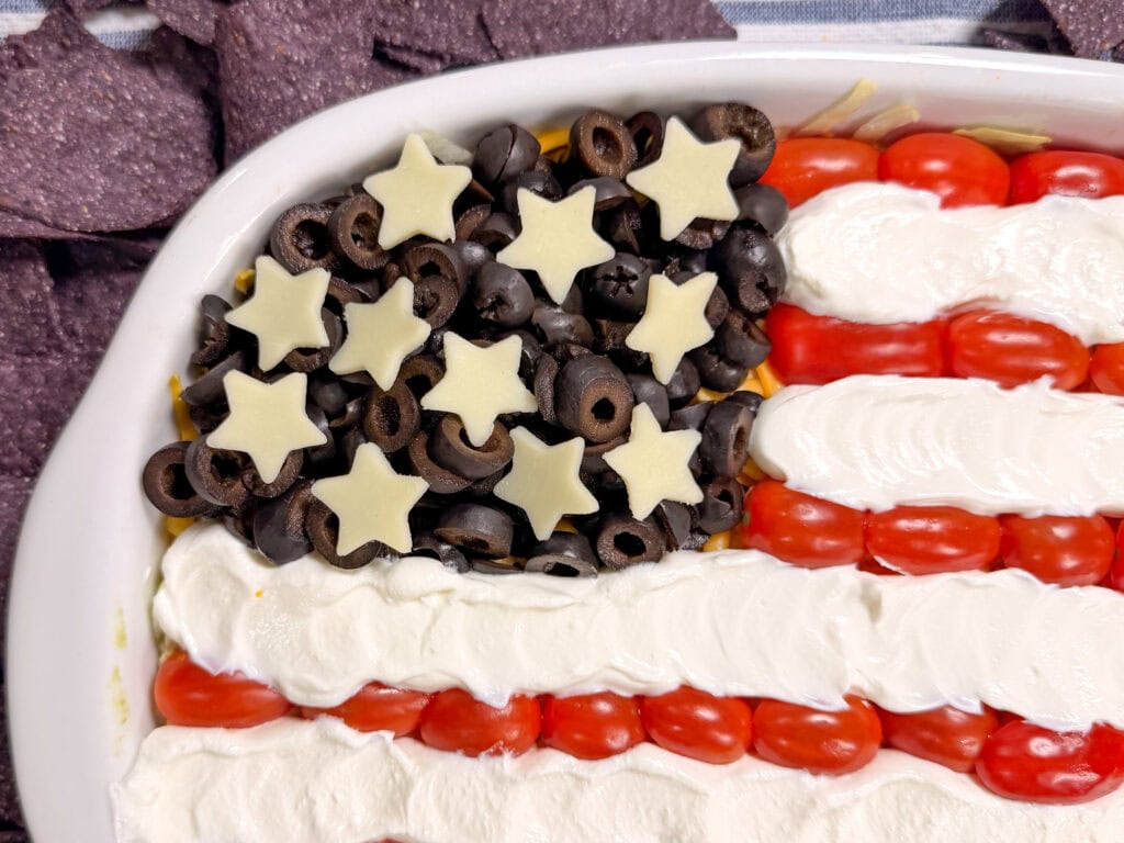 A layered dip in a dish designed to look like the American flag, using sliced black olives and star-shaped cheese for the stars, cherry tomatoes for the stripes, and a creamy white topping for the white stripes.