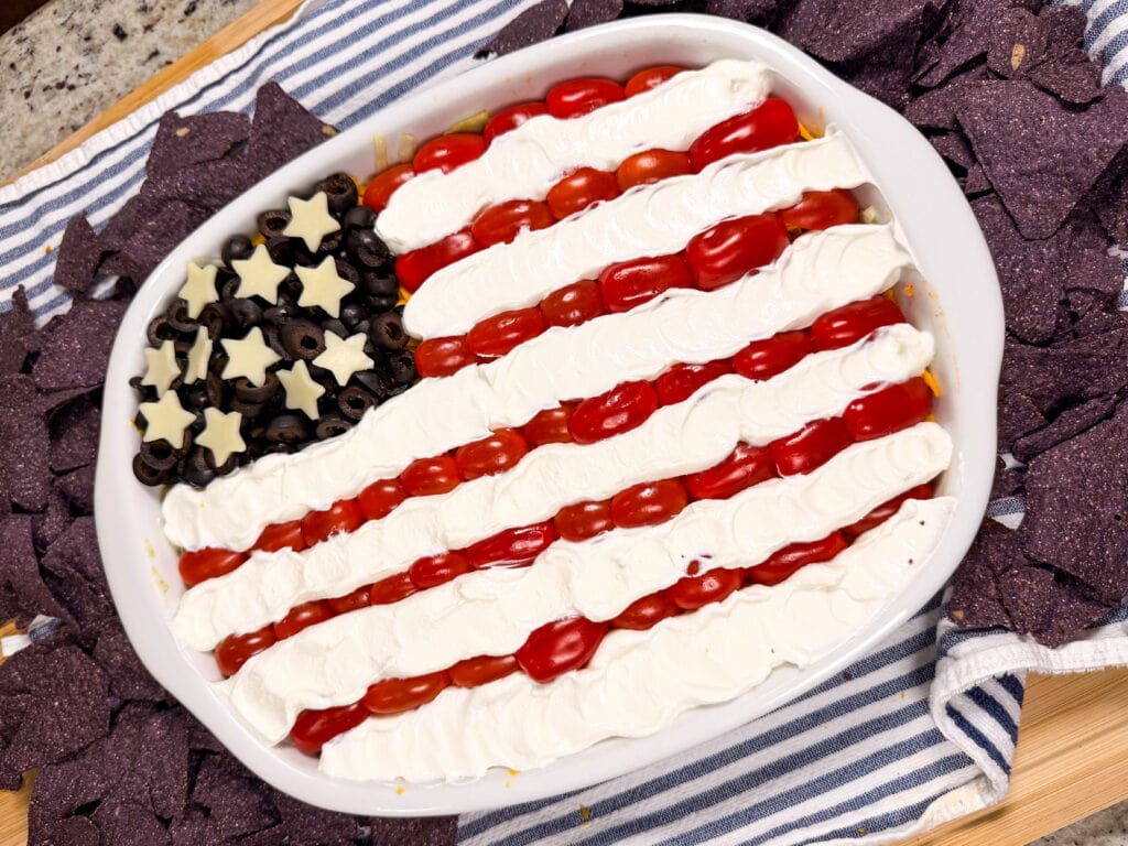 A dip styled like the American flag, with grape tomatoes and sour cream for stripes, sliced black olives and star-shaped cheese for the stars, served in a white dish surrounded by blue corn tortilla chips.