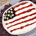 A dip styled like the American flag, with grape tomatoes and sour cream for stripes, sliced black olives and star-shaped cheese for the stars, served in a white dish surrounded by blue corn tortilla chips.