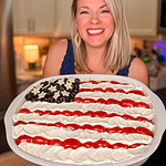 A smiling woman in a kitchen holds a large platter decorated like the American flag with layered dip, using black olives for stars, and rows of sour cream and salsa for stripes.