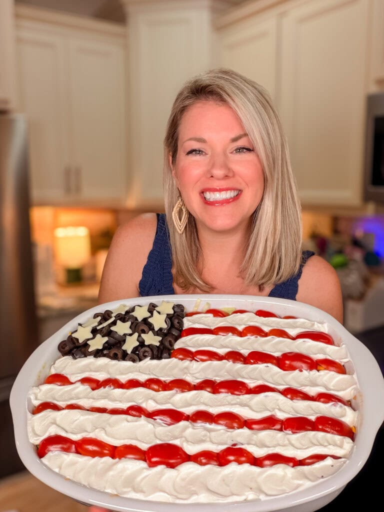 A smiling woman in a kitchen holds a large platter decorated like the American flag with layered dip, using black olives for stars, and rows of sour cream and salsa for stripes.